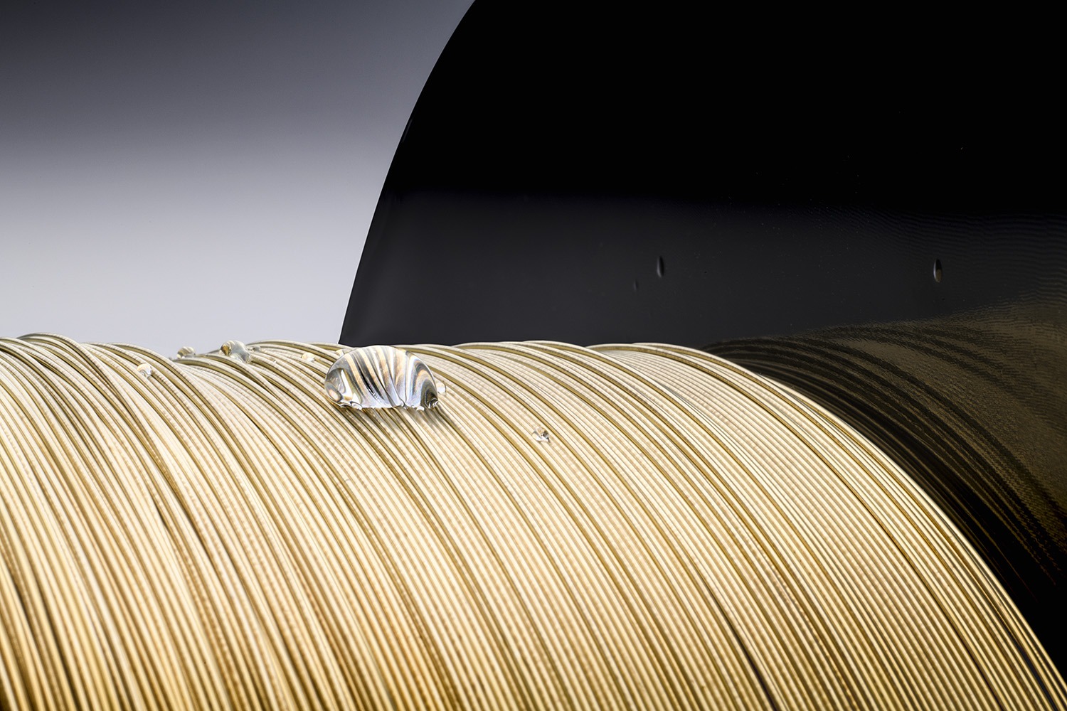 Close-up of a spool of shiny, golden metal wire with a water droplet resting on it, set against a dark background, emphasizing the wire's texture and shine.