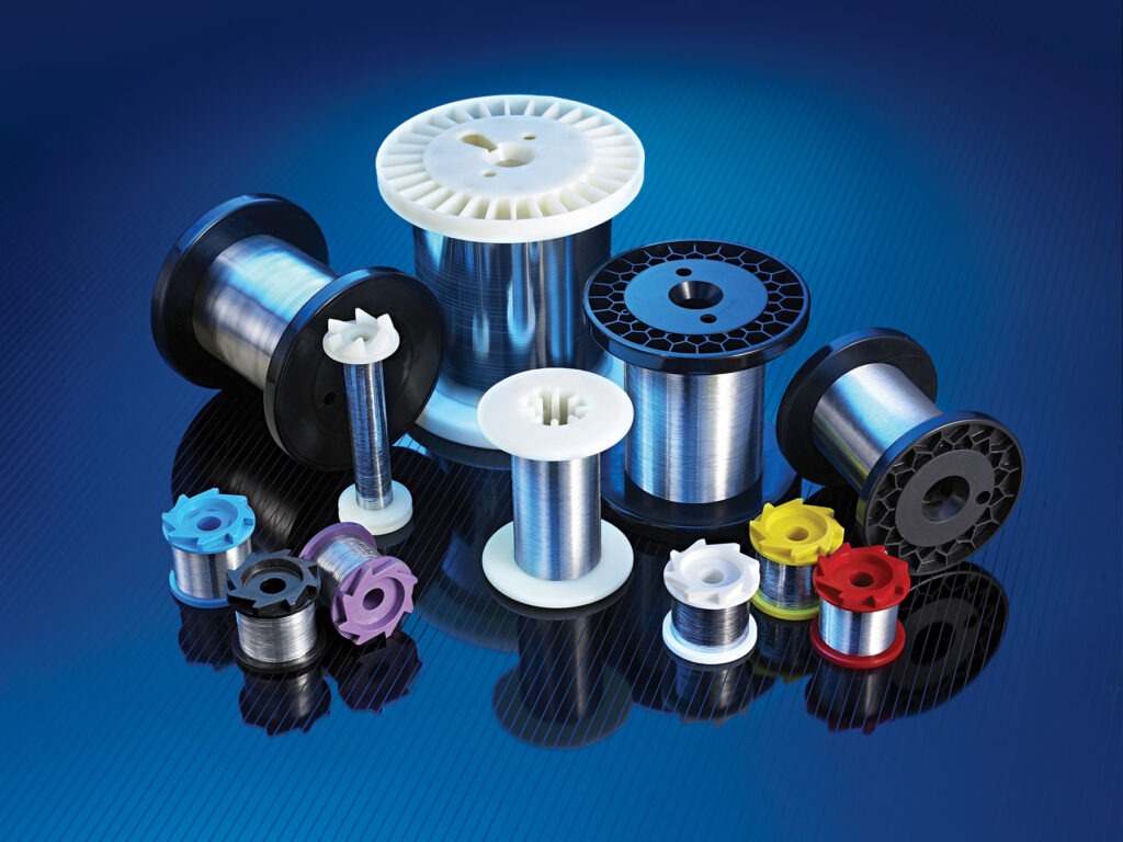 Various spools of metallic wire in different colors and sizes arranged on a reflective blue background. Suitable for manufacturing and crafting purposes.