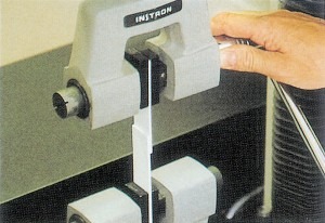 Material testing apparatus grips a specimen while a hand adjusts the setup, demonstrating tension or compression testing for material properties.